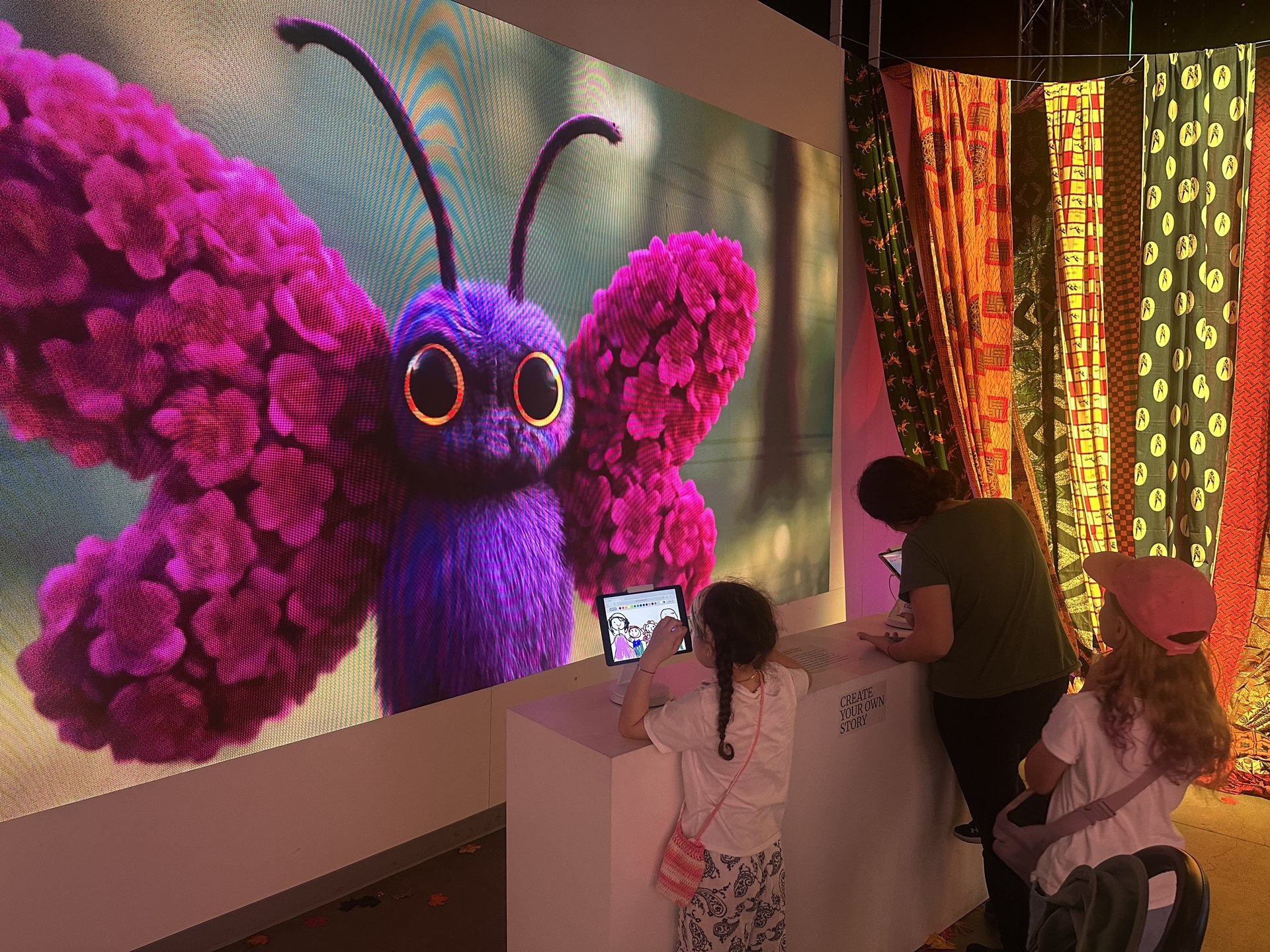 A child draws on a tablet beside a giant purple butterfly and other guests in Mythos