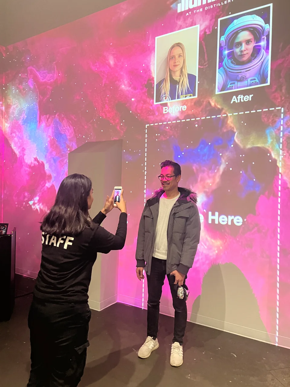 A staff member photographs a smiling guest beneath a projected before-and-after astronaut portrait at Illuminarium