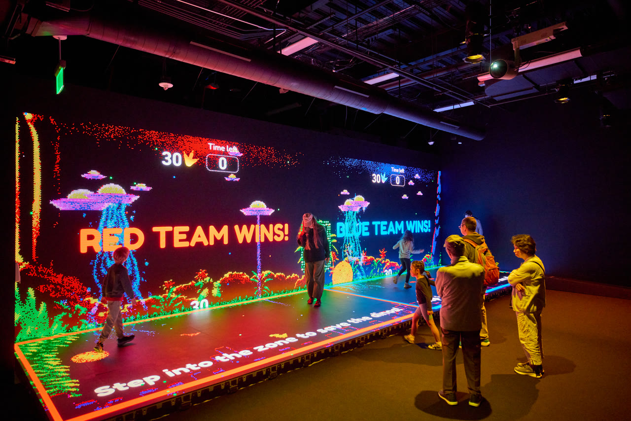 Visitors on an illuminated play platform in front of a giant Lite-Brite arcade wall showing red and blue team results